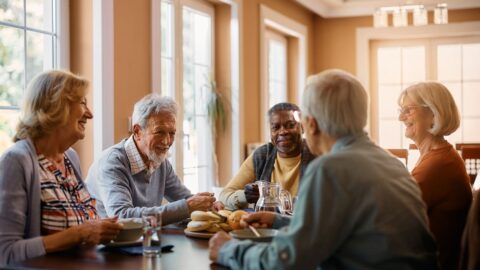 Group of senior people enjoying conversation and making new friends during lunch at dining table at senior living community