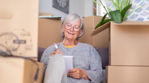 downsizing Happy senior woman involved in move to senior living sitting among cardboard boxes making note of purchases and to-dos, concept of moving, retirement, downsizing, new life, buying, renting, apartment, house