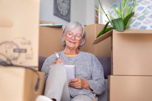 Happy senior woman involved in move to senior living sitting among cardboard boxes making note of purchases and to-dos, concept of moving, retirement, new life, buying, renting, apartment, house